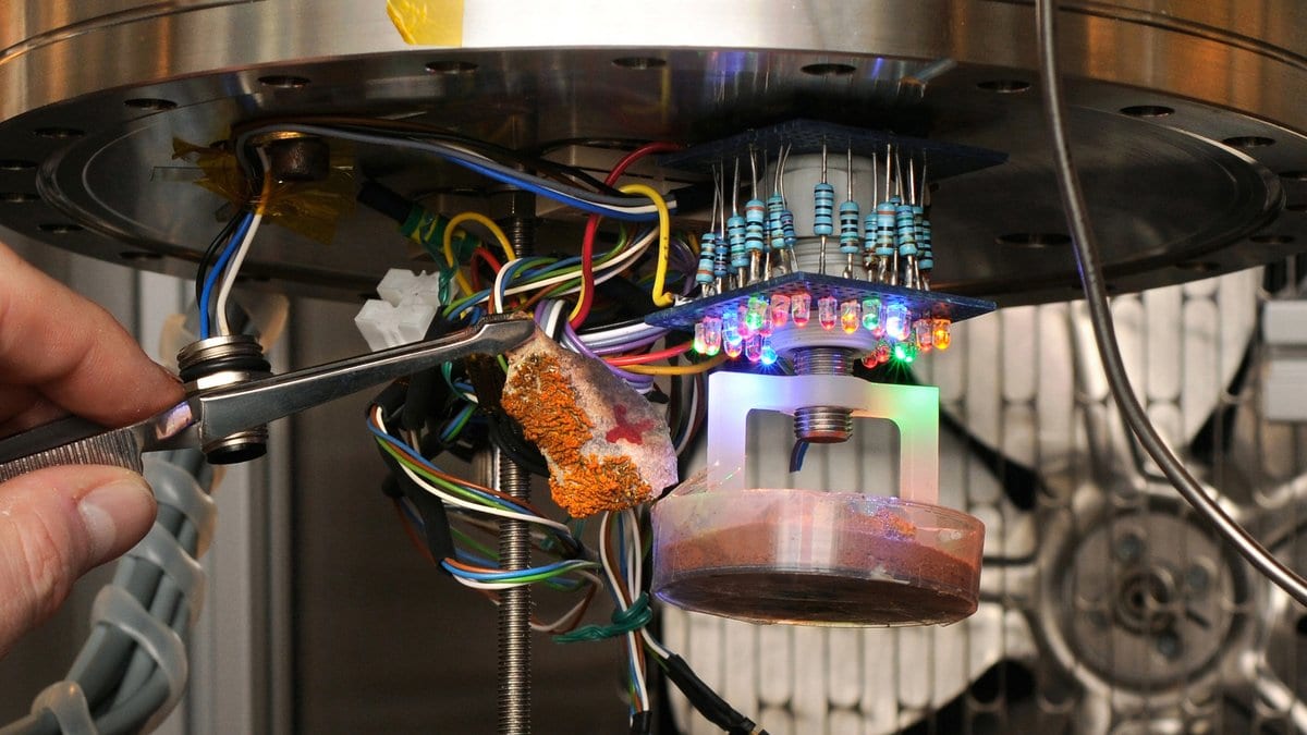 A gloved hand uses tweezers to place a rock sample inside a metal planetary simulation chamber ringed with lamps and wiring.