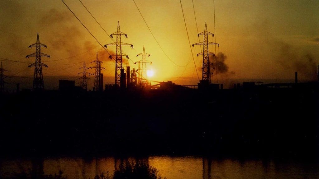 Blast furnaces and chimneys of a steel plant in silhouette behind high-voltage transmission towers, with smoke plumes rising into an orange sky.