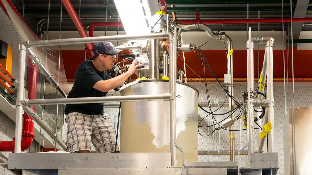 A physicist leans over the top of a tall metal trap assembly surrounded by pipes and cabling in a laboratory hall.