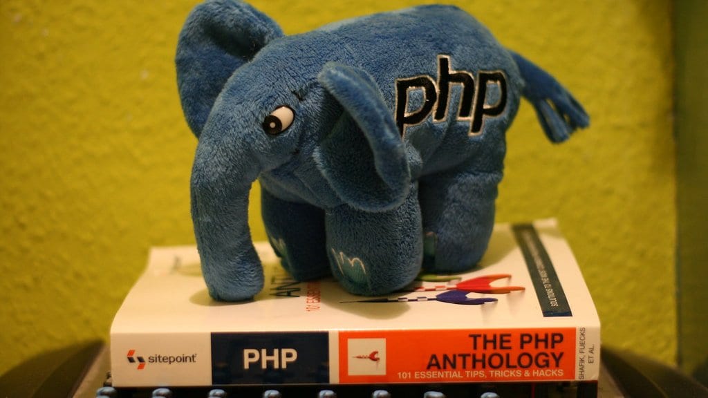A blue plush elephant with the word php on its side, standing on a PHP programming book.