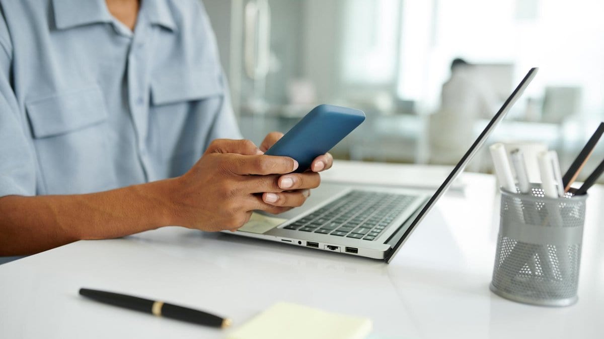 A person at an office desk holds a smartphone in one hand beside an open laptop.