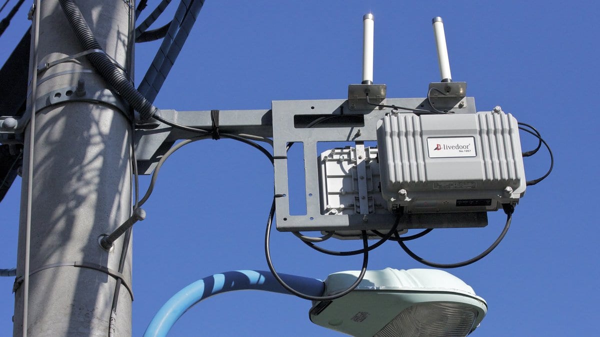 A gray weatherproof radio unit with two upright antennas, bolted to a bracket on a utility pole above a street lamp, against a clear blue sky.