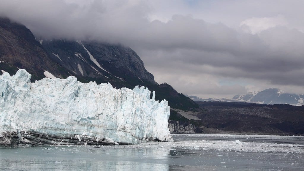 A glacier's ice cliff ends in seawater in Glacier Bay, Alaska, with small icebergs and brash ice drifting in front of it below cloud-covered mountains.