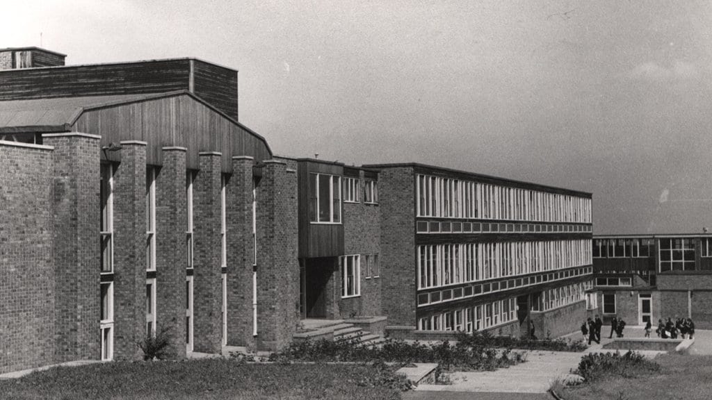 A long, low brick-and-glass secondary school building of the 1960s seen from a path across grass, with a small group of pupils gathered near the entrance.