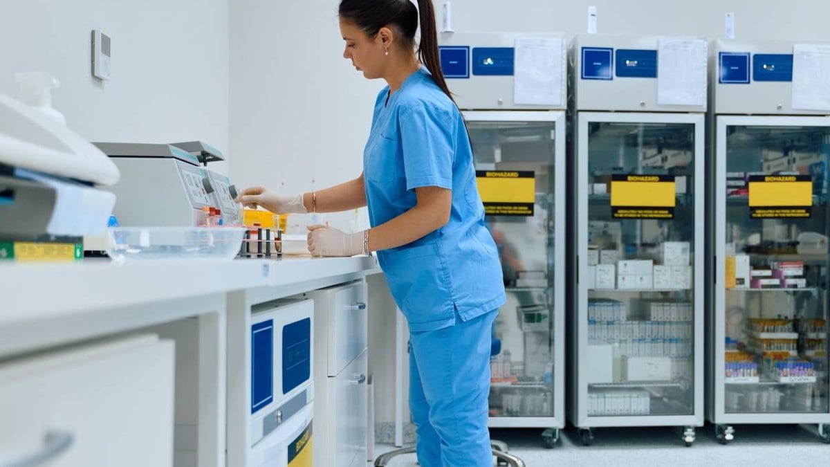 A laboratory technician in blue scrubs places blood sample tubes into an automated analyzer, with refrigerated storage cabinets behind her.
