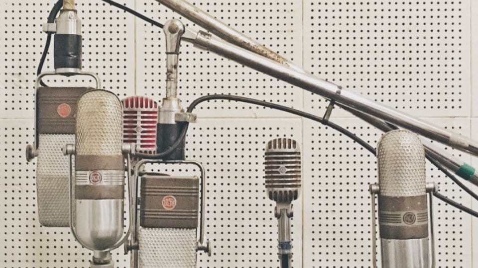 A bank of vintage studio microphones on stands and a boom arm in front of a perforated acoustic wall.