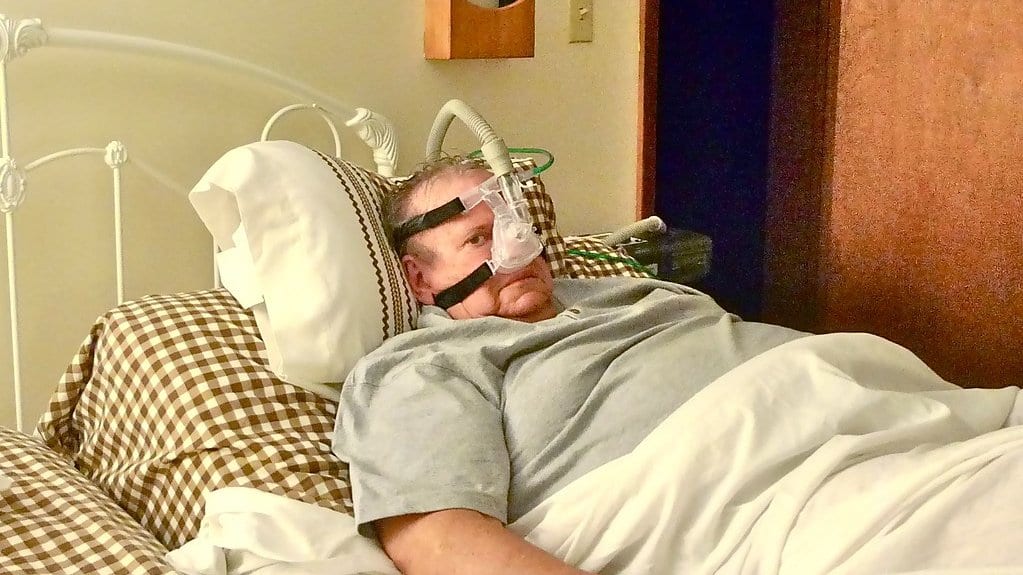 A man lies in bed wearing a nasal mask connected by a flexible hose to a bedside machine that delivers pressurized air.