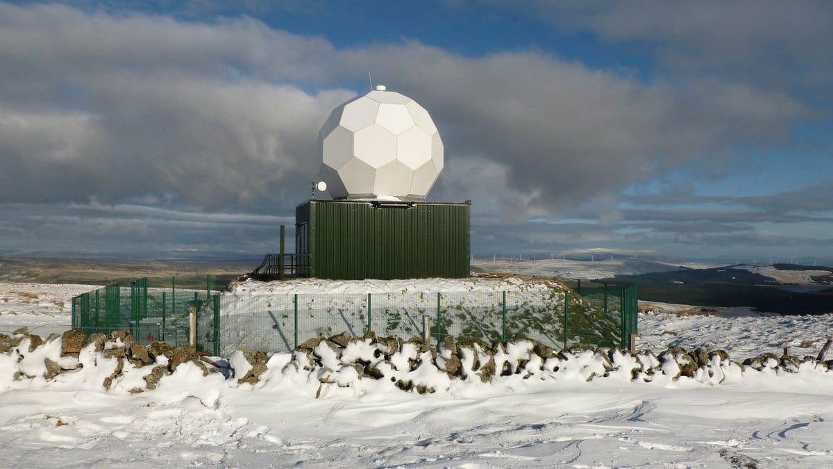 A white geodesic radar dome on a green cabin inside a fenced compound on a snowy hilltop, a drystone wall drifted with snow in front