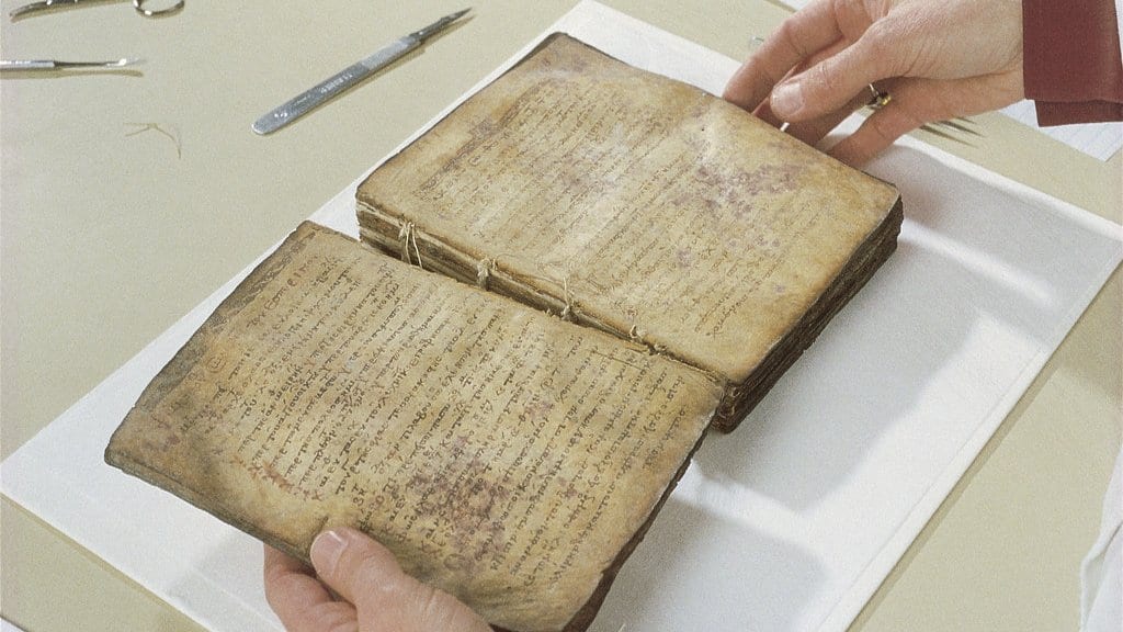 Hands hold an open medieval codex of worn parchment on a conservation table, with small tools beside it.
