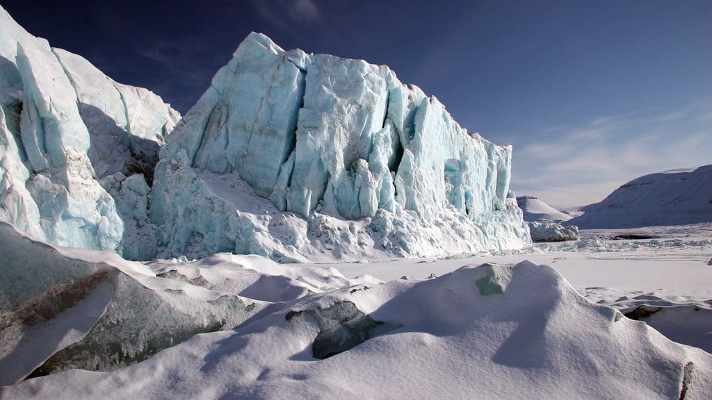 A wall of pale blue glacier ice above snow-covered ground in Svalbard, with low dark hills on the horizon.