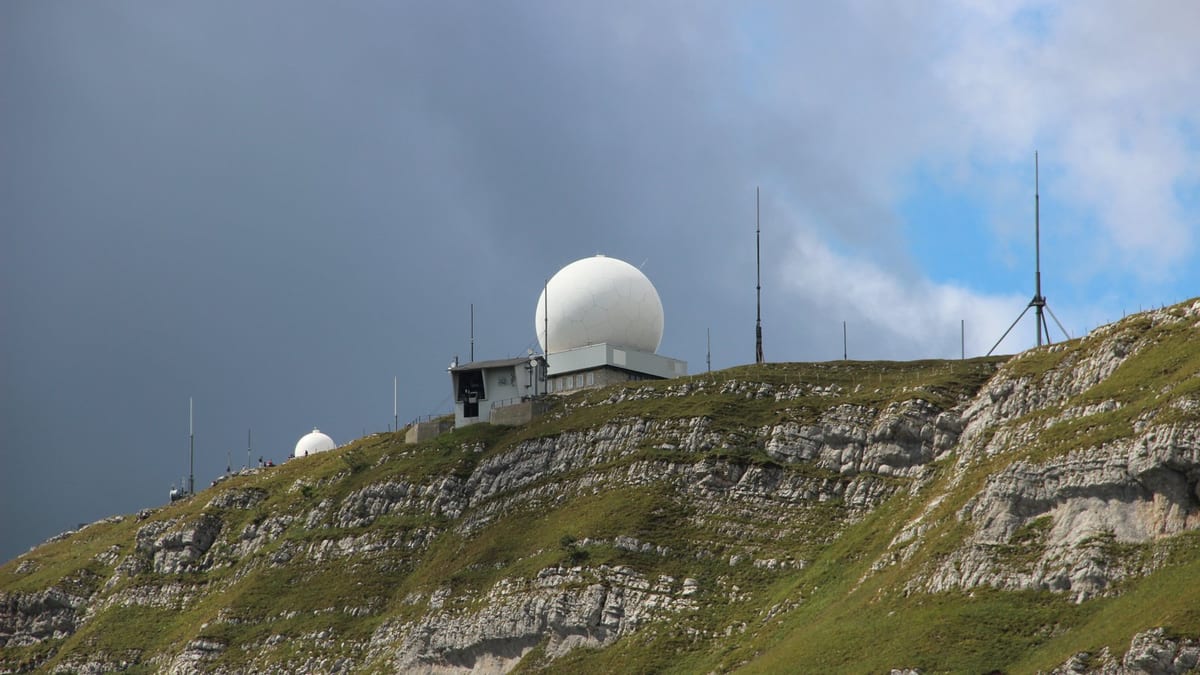 A white spherical radar dome on a small building at the top of a grassy limestone ridge, dark storm cloud filling the sky behind, masts along the crest