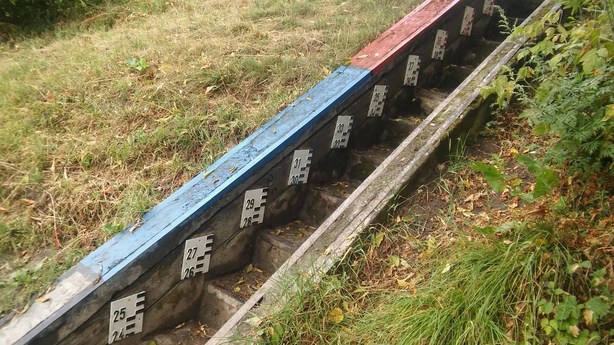 Numbered metal gauge plates fixed in steps down a long wooden frame painted blue and red, running diagonally through grass and weeds