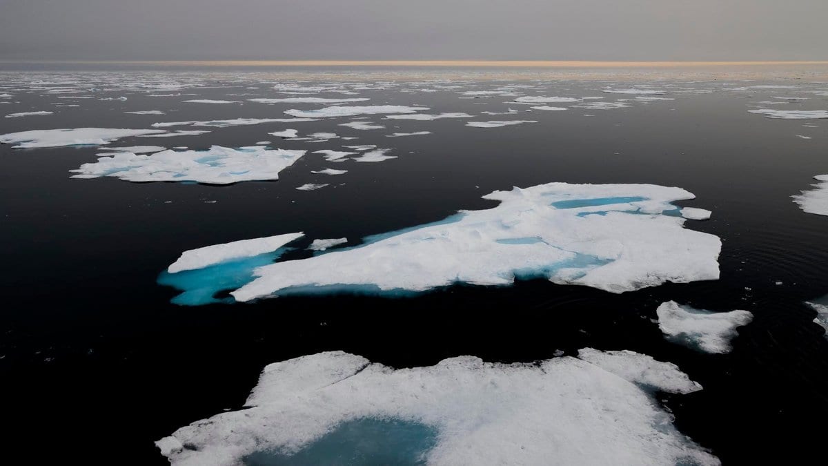 Floating sea ice floes with pale blue meltwater pools, separated by dark open ocean under a hazy sky.