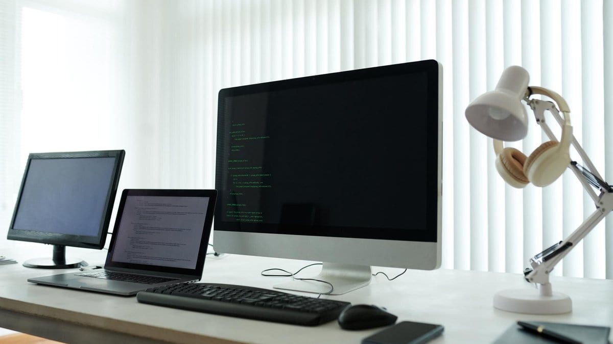 A desktop monitor showing lines of source code on a dark background, beside a laptop and a keyboard on a white desk in a bright office.
