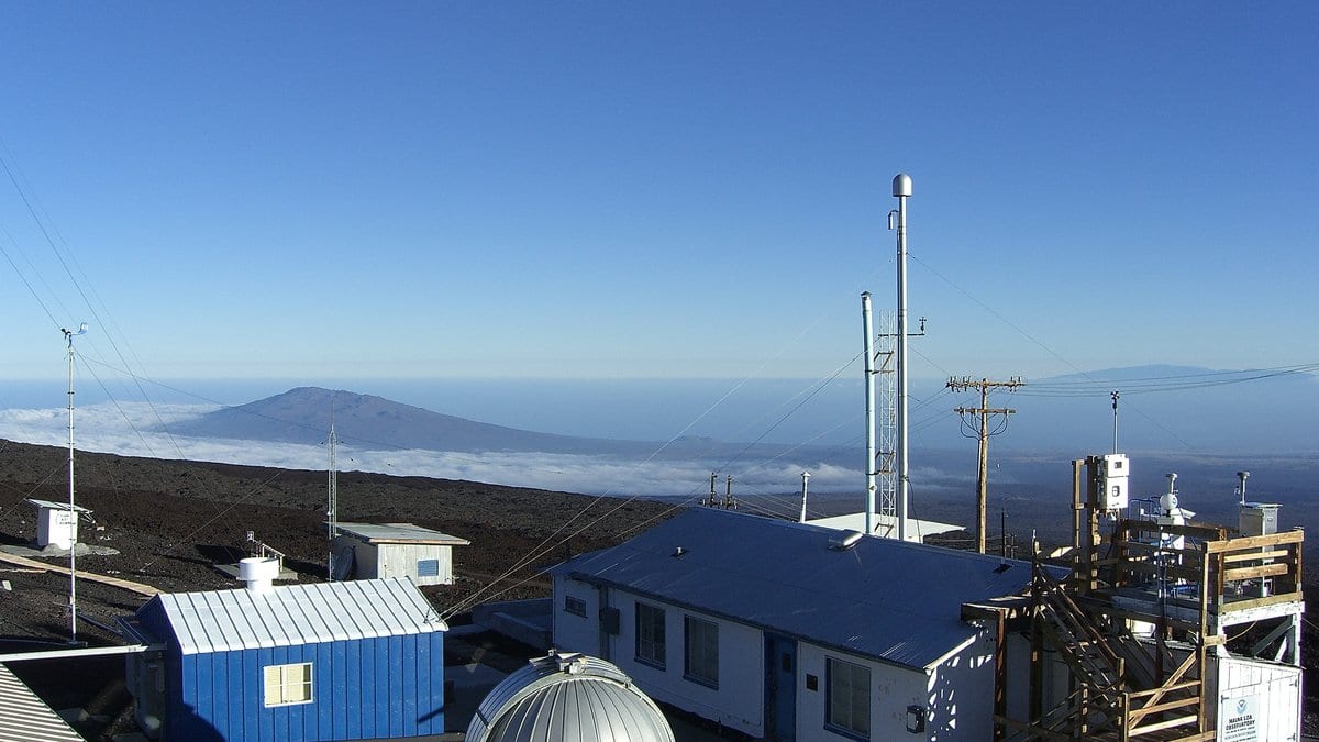 The Mauna Loa Observatory in Hawaii, its air-sampling masts rising above a cloud deck on a bare lava slope.