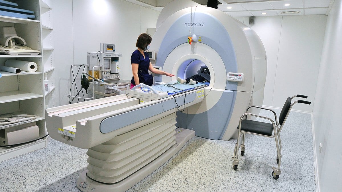 A technologist stands beside a magnetic resonance imaging scanner in a clinical imaging room, with the patient table drawn out in front of the open bore.