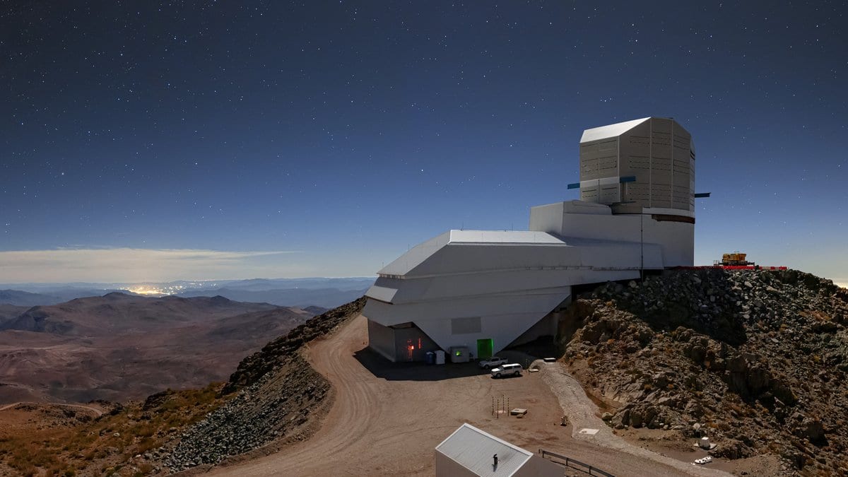 The Vera C. Rubin Observatory building on a mountain ridge at night, stars overhead and distant valleys below.