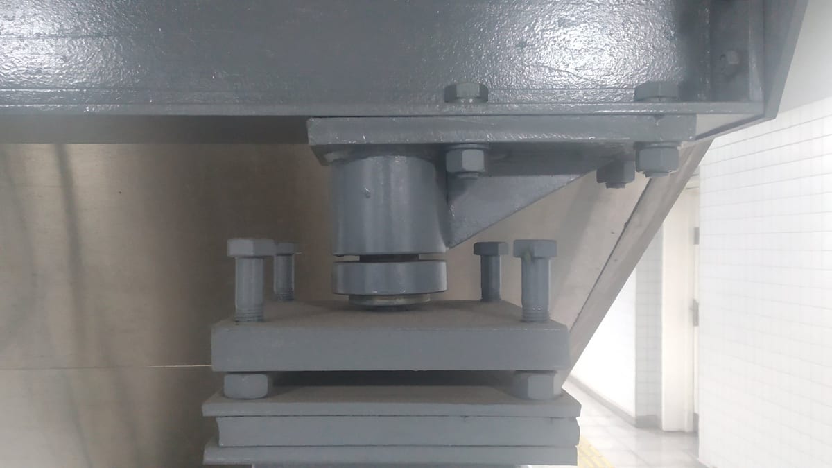 A grey-painted steel bearing assembly bolted between a heavy beam and a stack of steel plates, a white tiled wall to one side
