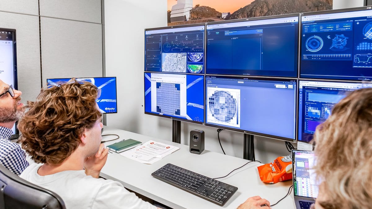 Three researchers sit at a desk in a laboratory control room, facing a wall of monitors showing control-system dashboards and sky images.