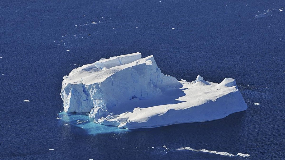 An aerial view of a flat-topped iceberg and scattered ice floes in dark blue water off Antarctica.