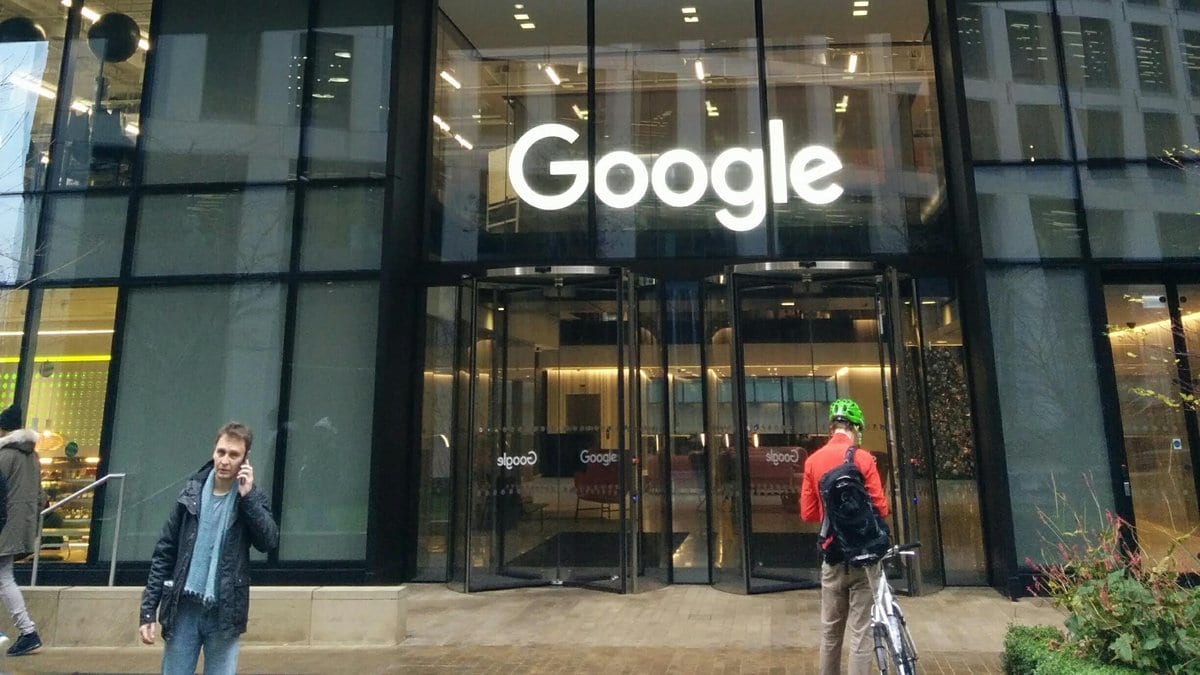 The lit Google sign above the glass entrance of the company office building at 6 Pancras Square in London, with people walking past on the sidewalk.