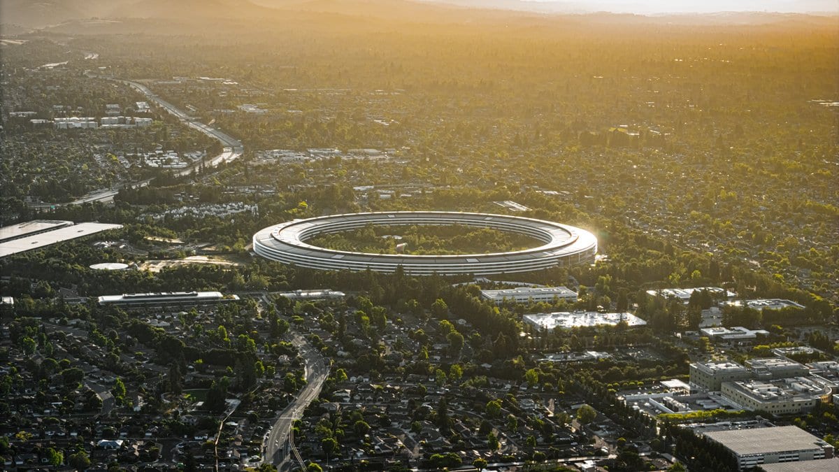 Aerial view at sunset of Apple Park, the ring shaped headquarters building of Apple in Cupertino, California, surrounded by trees and suburban streets.