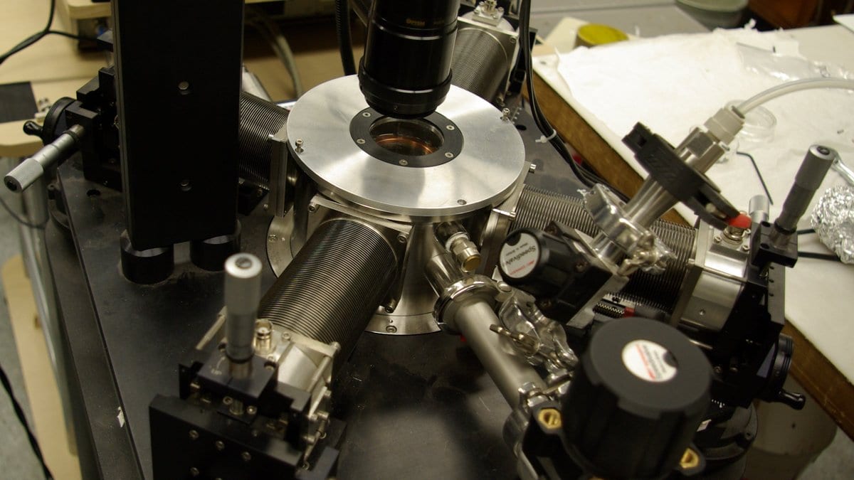 A cryogenic probe station on a laboratory bench, with a small vacuum chamber at its center and several micromanipulator arms carrying fine electrical probes toward the sample inside.