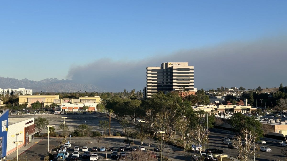 Gray wildfire smoke stretching across the sky above a San Fernando Valley shopping center, with mountains in the distance.