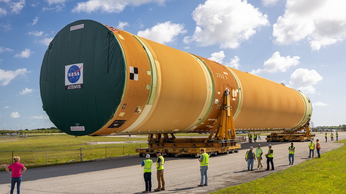 A long orange rocket stage on a wheeled transporter moves along a road under a blue sky, with workers in high-visibility vests watching from the roadside.