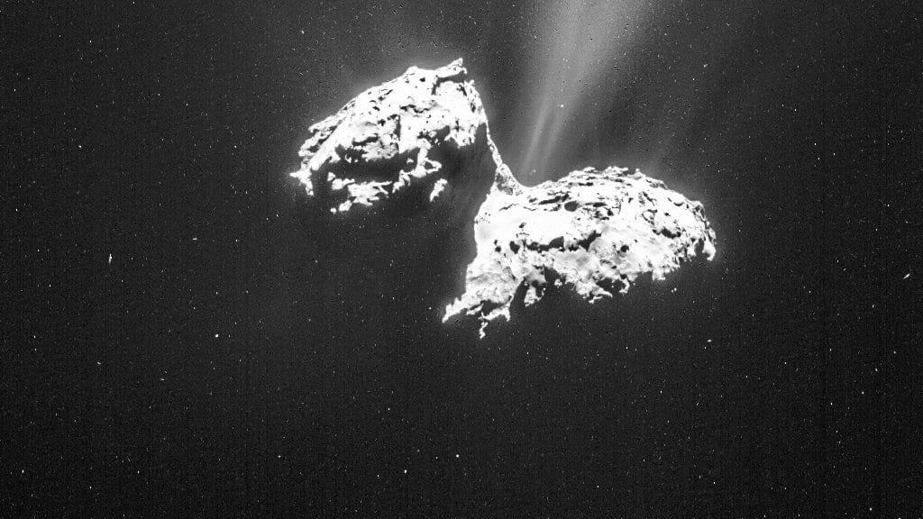The two lobed icy nucleus of comet 67P/Churyumov-Gerasimenko, lit from the left, with faint dust jets rising from the neck between its lobes against a black sky.