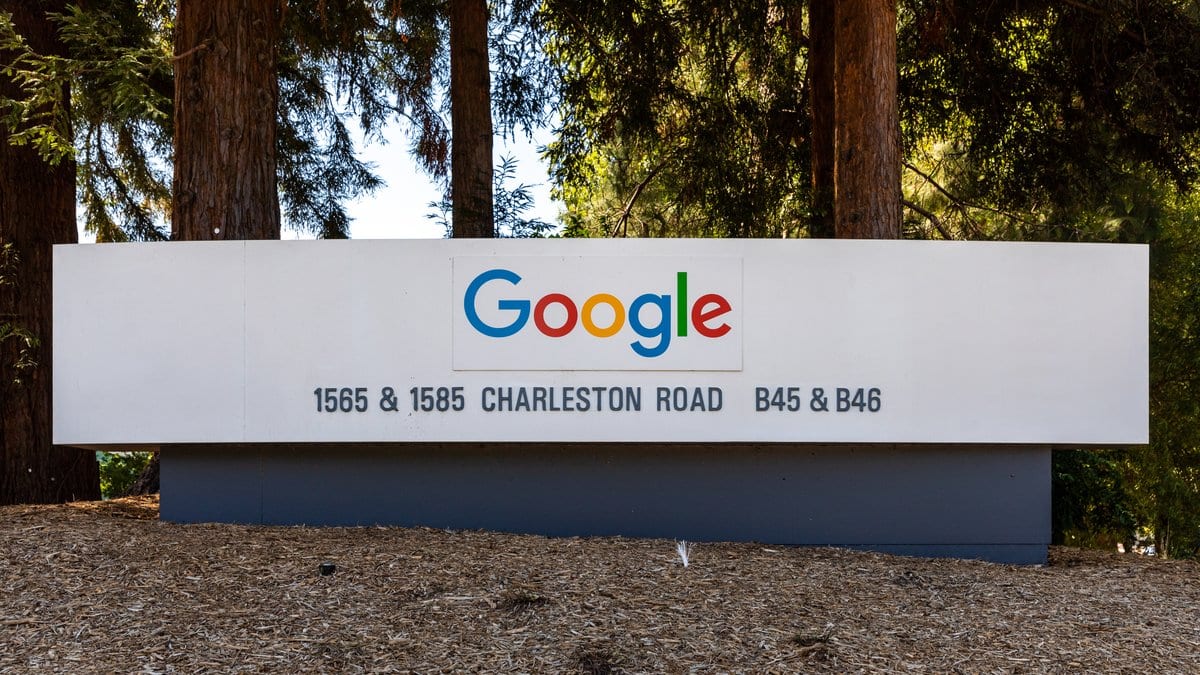 A white sign bearing the multicolor Google logo stands among tall trees at the entrance to a company campus.