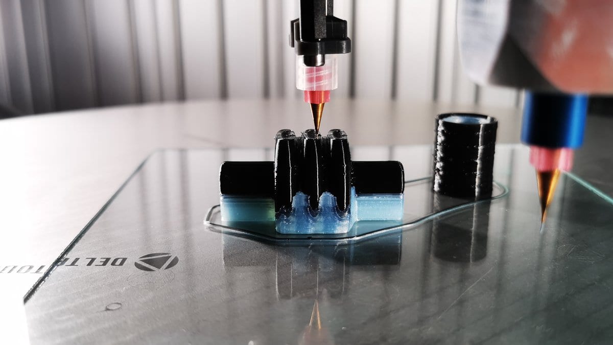 A 3D printer nozzle extrudes black silicone elastomer onto a glass build plate, with blue support material around the printed part.