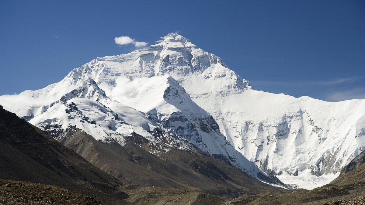 Mount Everest's snow covered north face rising above a brown rocky valley under a deep blue sky.