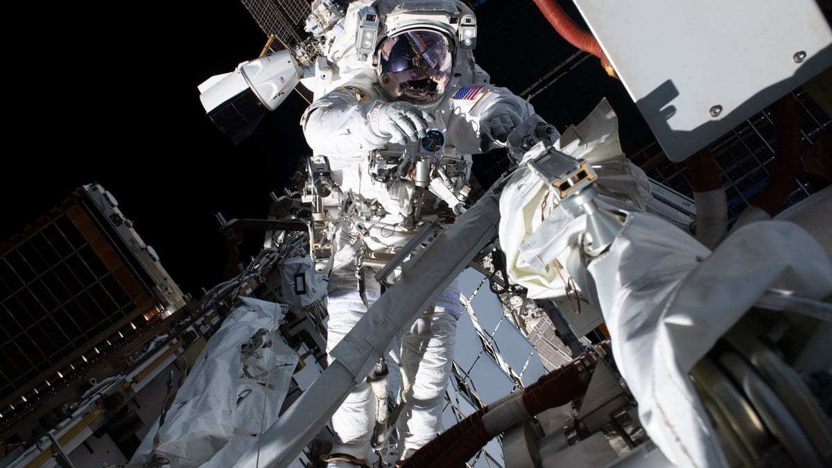 A spacewalker in a white spacesuit holds onto the International Space Station truss, lit against black space.