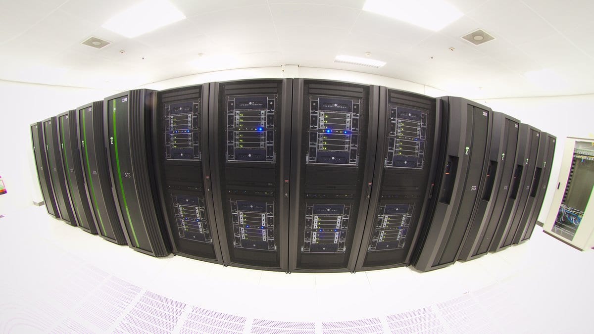 A wide view of rows of tall black computing cabinets in a brightly lit machine hall.