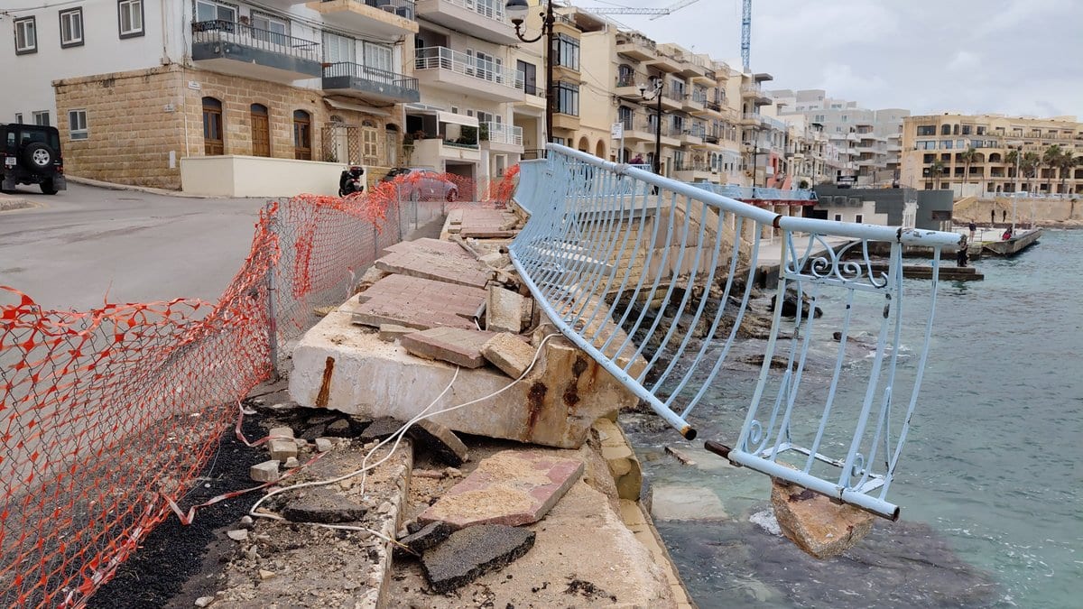 A seafront promenade with its metal railing torn away and hanging over the water, and large paving slabs lifted from the walkway
