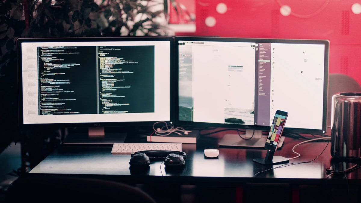 Two desktop monitors on a dark desk, the left screen filled with colored lines of code in an editor, with a keyboard, headphones and a phone in front of them.