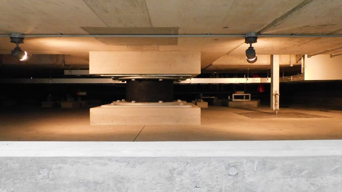 A single black cylindrical isolator under a concrete column head in a wide, low basement lit by spotlights, a concrete ledge across the foreground