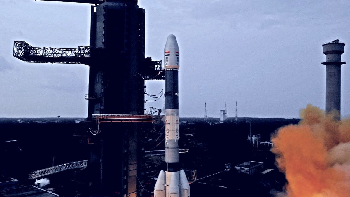 A GSLV Mk II rocket rises from its pad at Sriharikota, India, beside the mobile launch tower, with an orange exhaust cloud spreading across the launch complex.