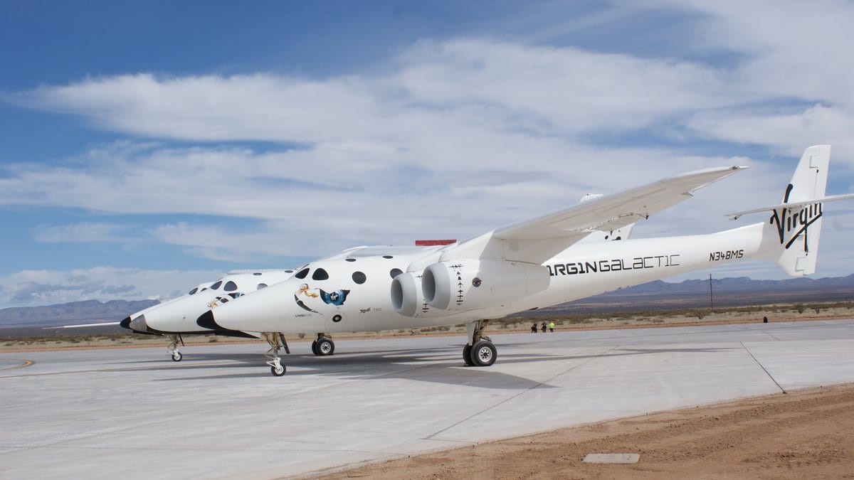 A white twin-fuselage carrier aircraft parked on concrete in the desert, with a small white spaceplane mounted between its two fuselages.