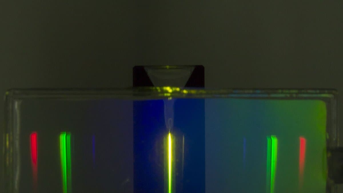 In a darkened laboratory, a glass cuvette glows along a narrow vertical yellow-green line where an infrared laser passes through a suspension of upconverting nanoparticles, with red, green and blue diffracted copies of the emission spread to either side.