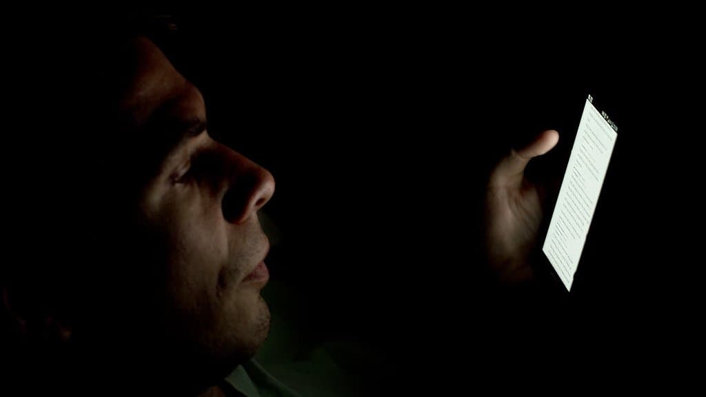 A man's face in profile, lit only by the screen of the phone he is reading in a dark room.