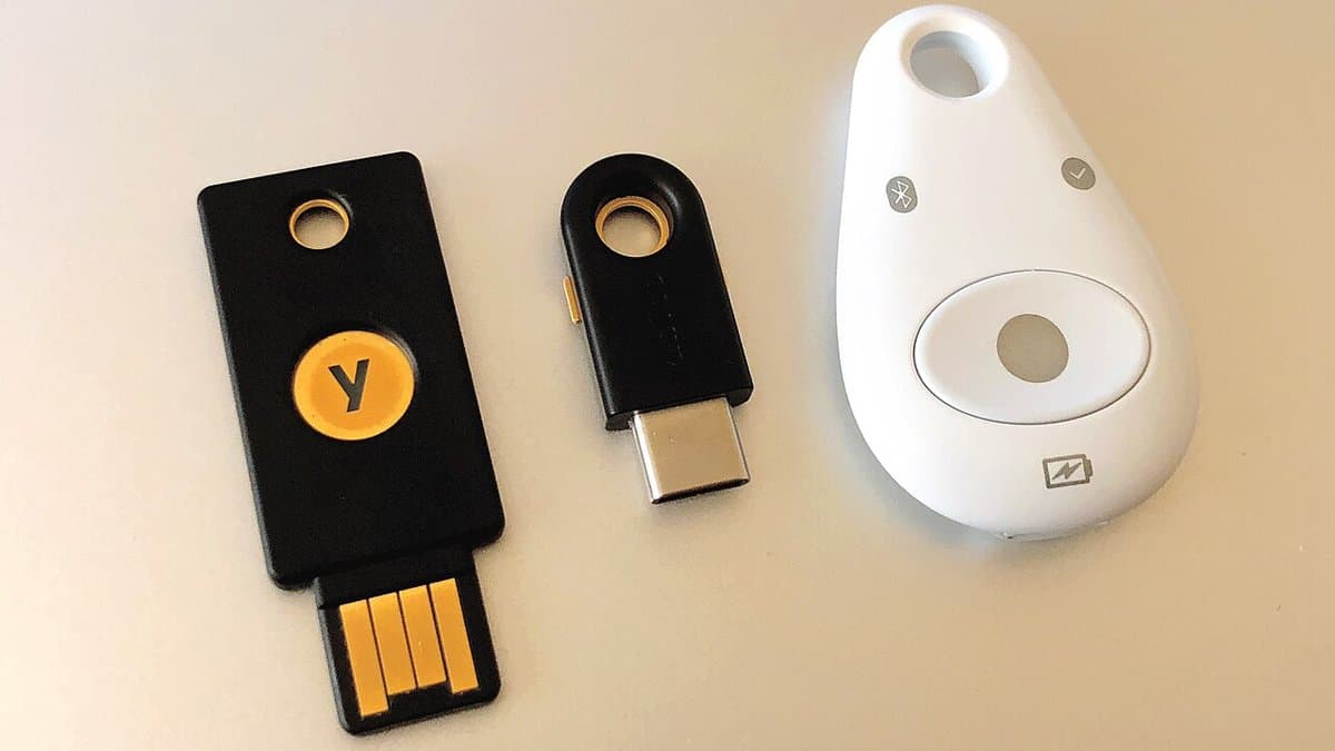 Three hardware authentication keys lying on a pale desk: two small black USB security keys and a rounded white Bluetooth key.