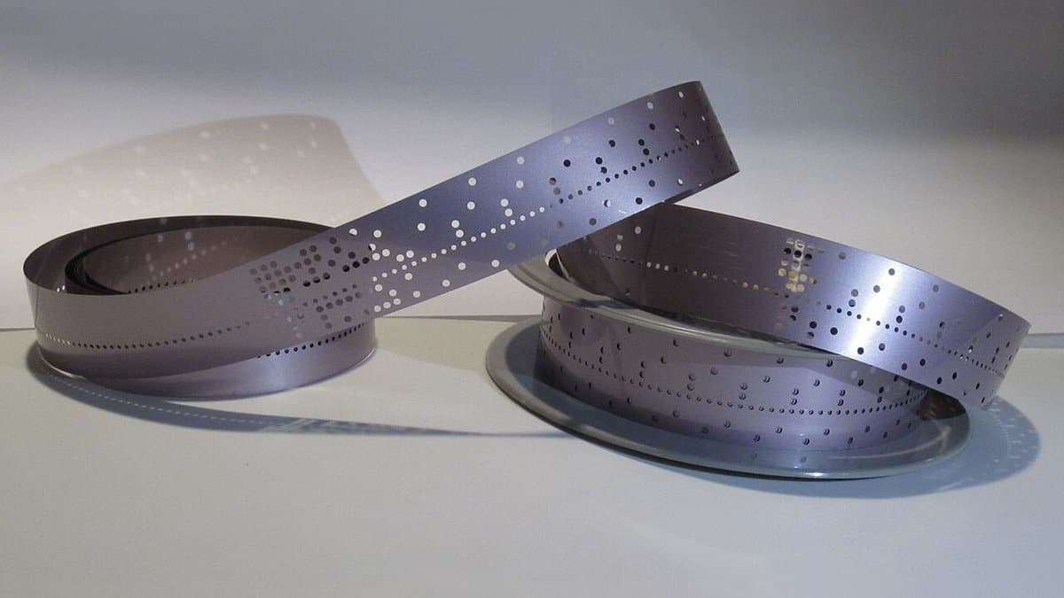 Two coils of silvery mylar punched tape on a pale surface, rows of small holes running the length of the strip.