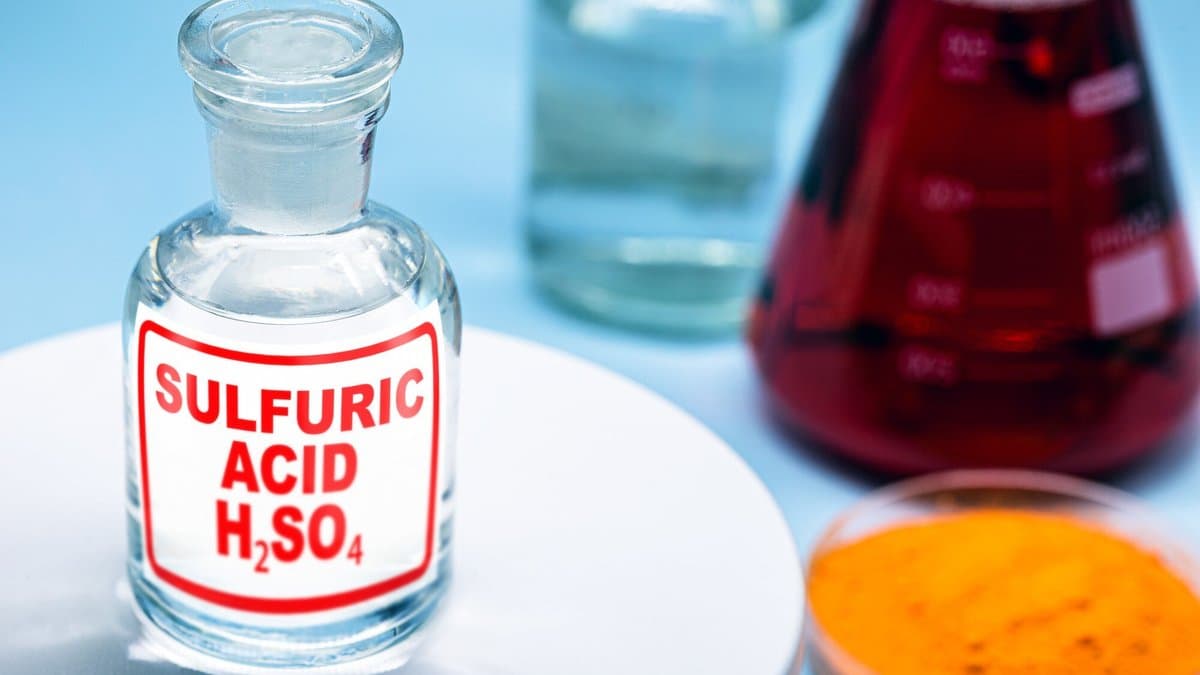 A glass reagent bottle labelled SULFURIC ACID H2SO4 stands on a white laboratory tray beside a flask of dark red liquid and a dish of orange powder.