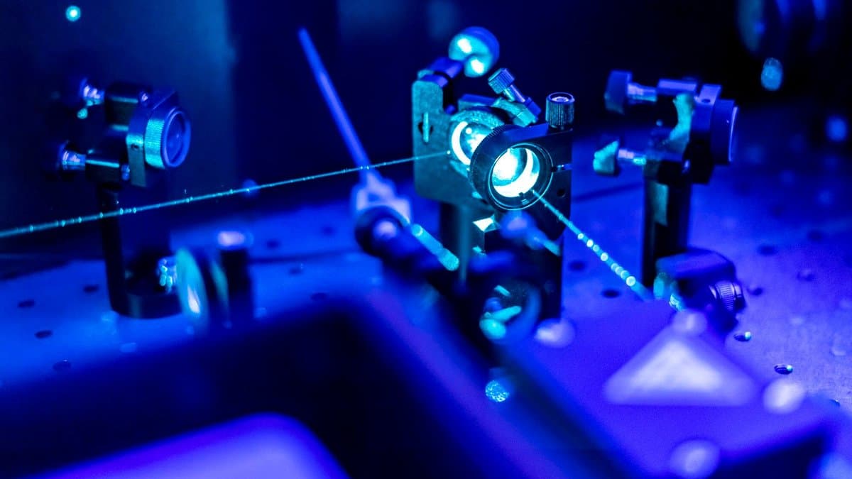 A blue laser beam crosses mirrors and lenses mounted on an optical table in a darkened laboratory.