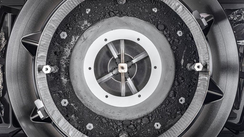 A circular sampler head seen from above, its inner ring packed with black asteroid dust and small rock fragments around a central collector.