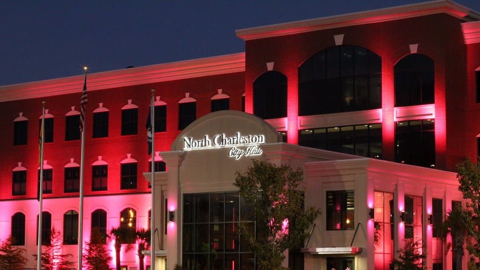 North Charleston City Hall at dusk, its brick facade washed in pink and red floodlights for Breast Cancer Awareness Month.