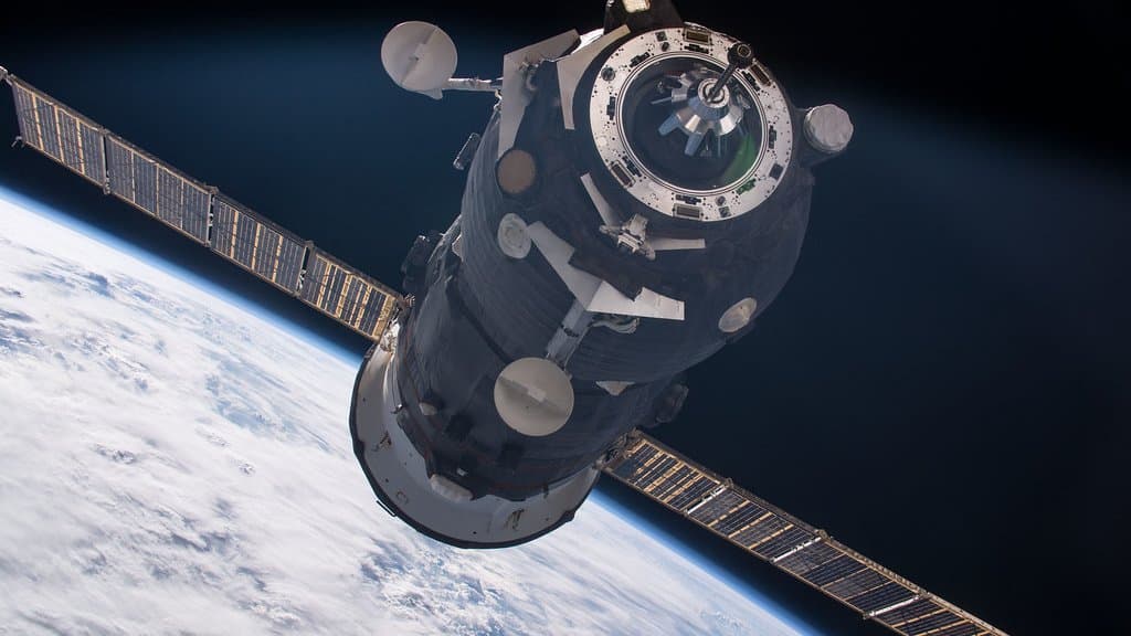 A Russian Progress cargo freighter in orbit above a cloud-covered Earth, its two solar arrays extended and its docking end facing the camera.