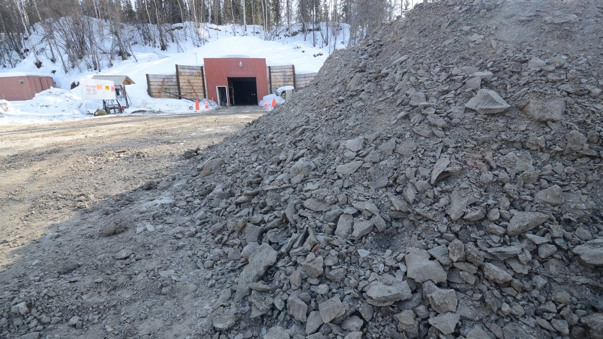 A tunnel portal set into a snowy hillside in a boreal forest, with a large pile of excavated gray frozen soil in the foreground.